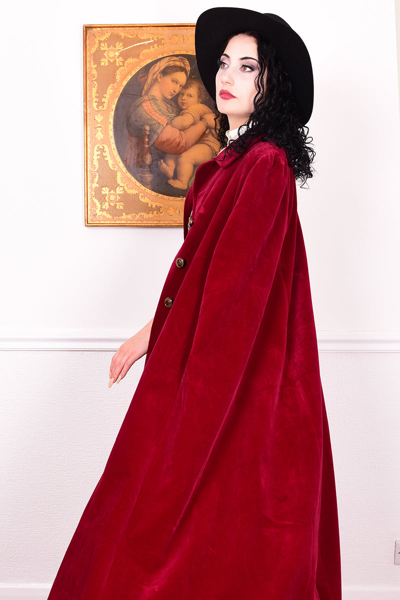 Woman in a vintage 1970s red velvet cape with a black hat standing in a room with a white wall.