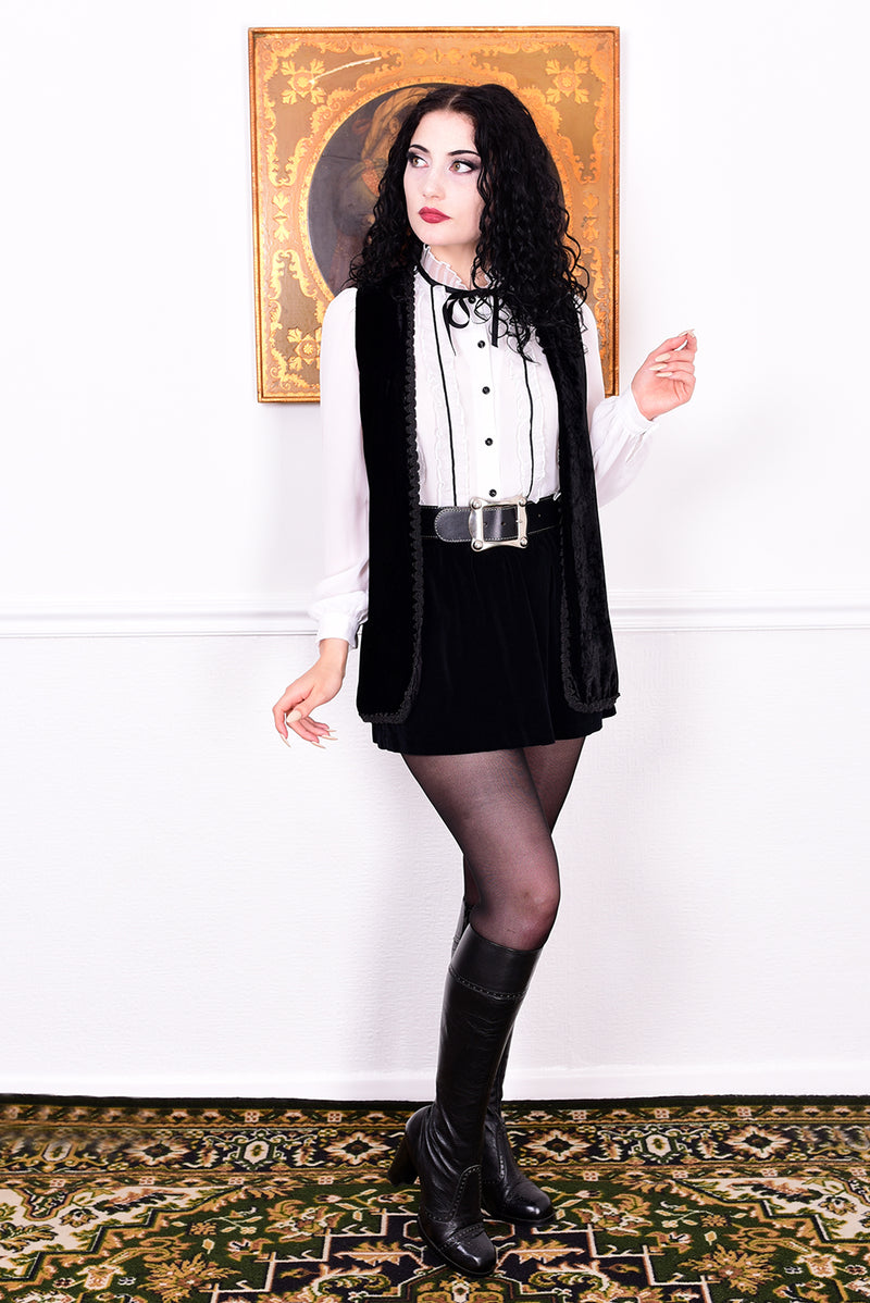 Woman in a black and white outfit with a belt and boots, standing in front of a white wall with a decorative rug.