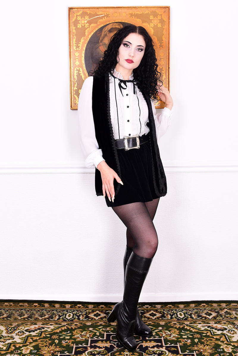 Woman in a black and white outfit with a belt and boots, standing in front of a decorative wall.