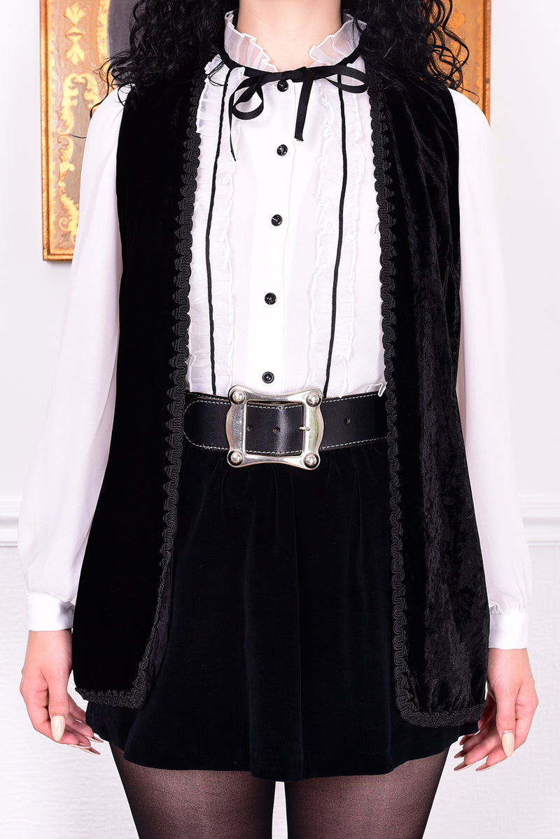 Person wearing a black vest over a white shirt with black buttons, standing against a plain background.