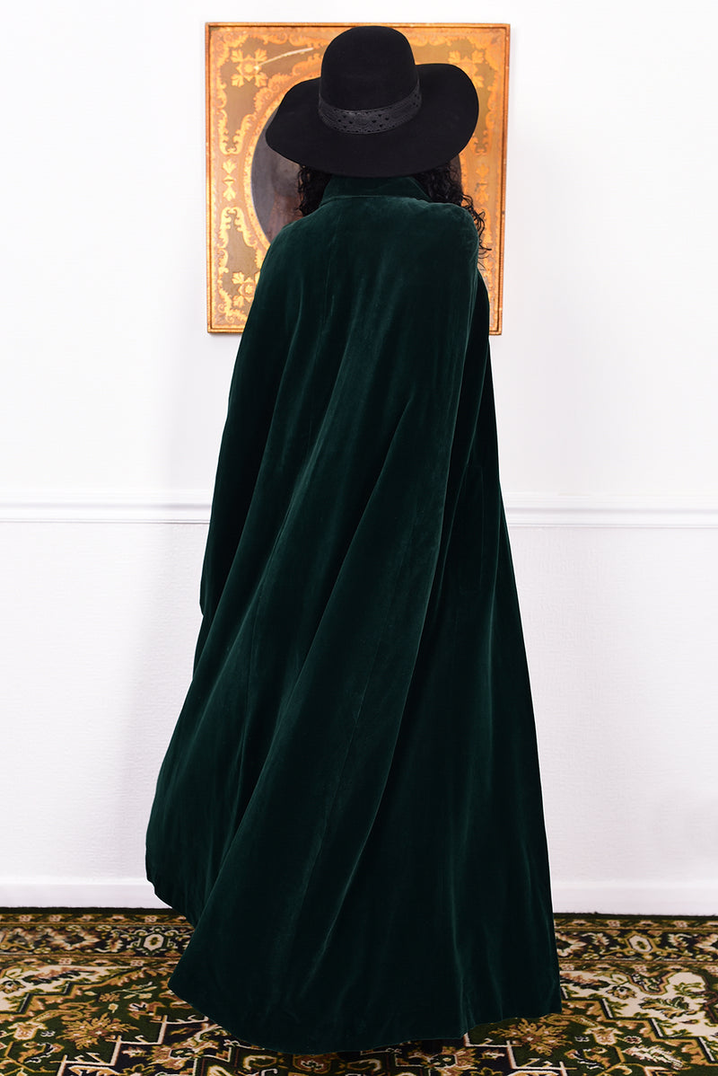 Woman wearing a vintage 1970s green velvet cape by Quad standing in a room with a white wall.