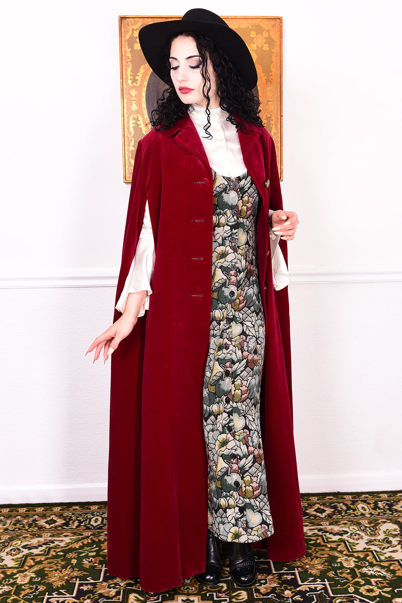 Woman in a vintage 1970s red velvet cape with a black hat standing in a room with a white wall.