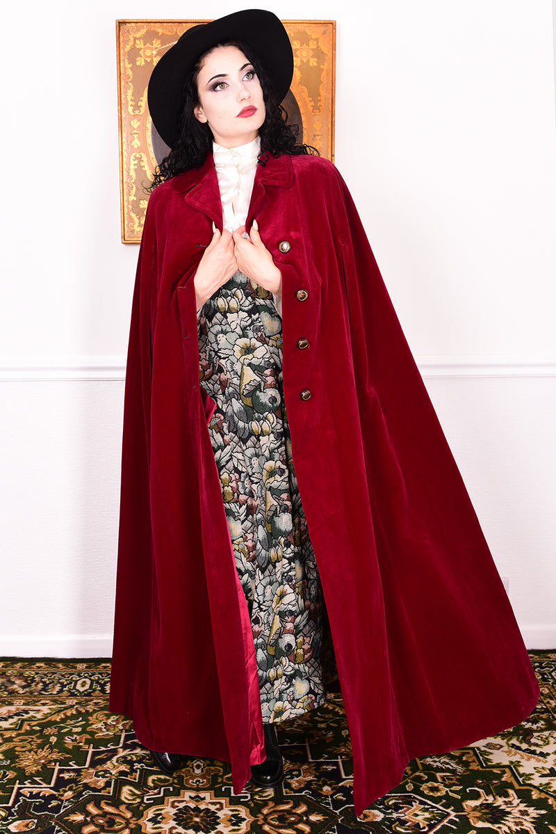 Woman in a vintage 1970s red velvet cape with a black hat standing in a room with a white wall.