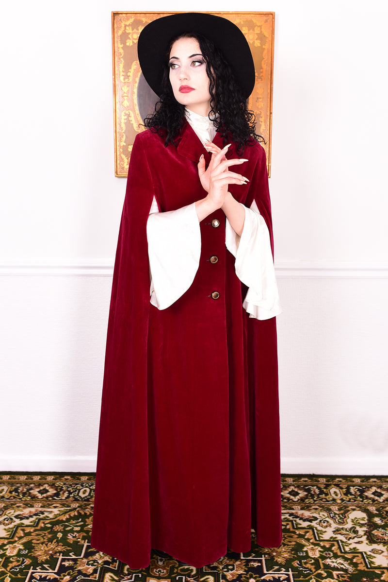 Woman in a vintage 1970s red velvet cape with a black hat standing in a room with a white wall.
