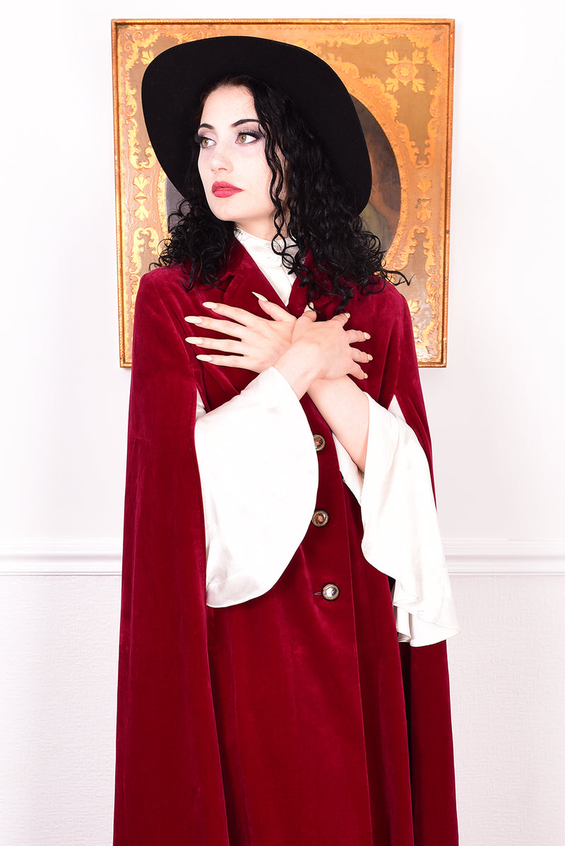 Woman in a vintage 1970s red velvet cape with a black hat standing in a room with a white wall.
