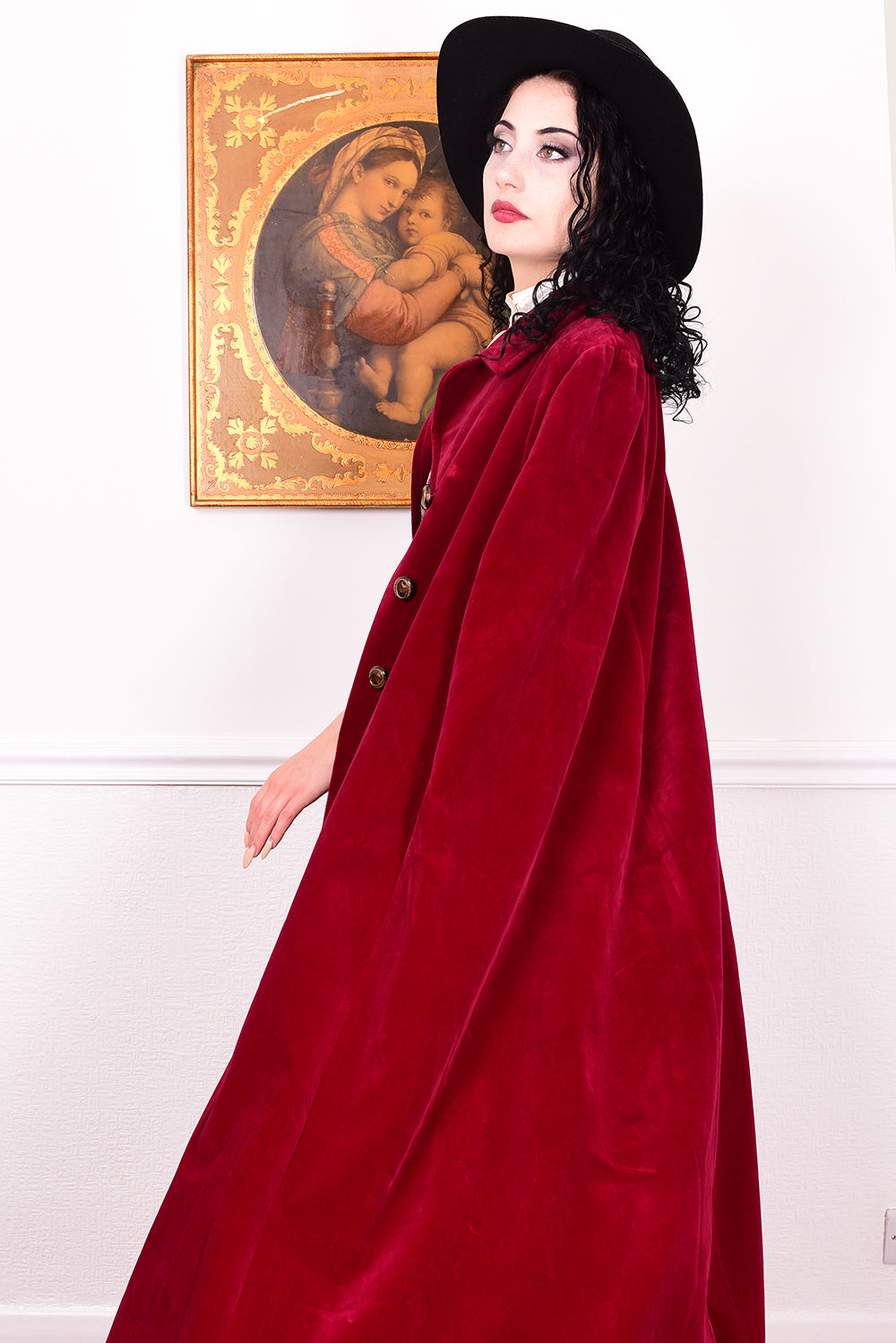 Woman in a vintage 1970s red velvet cape with a black hat standing in a room with a white wall.