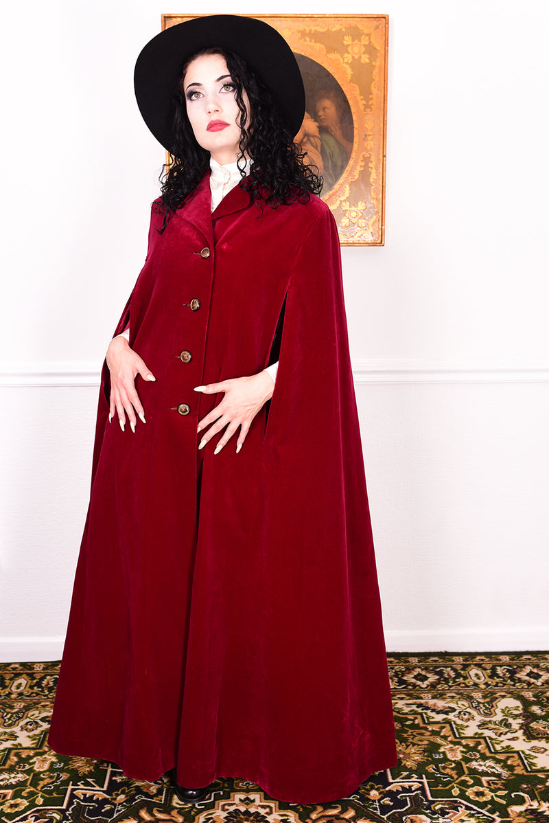 Woman in a vintage 1970s red velvet cape with a black hat standing in a room with a white wall.