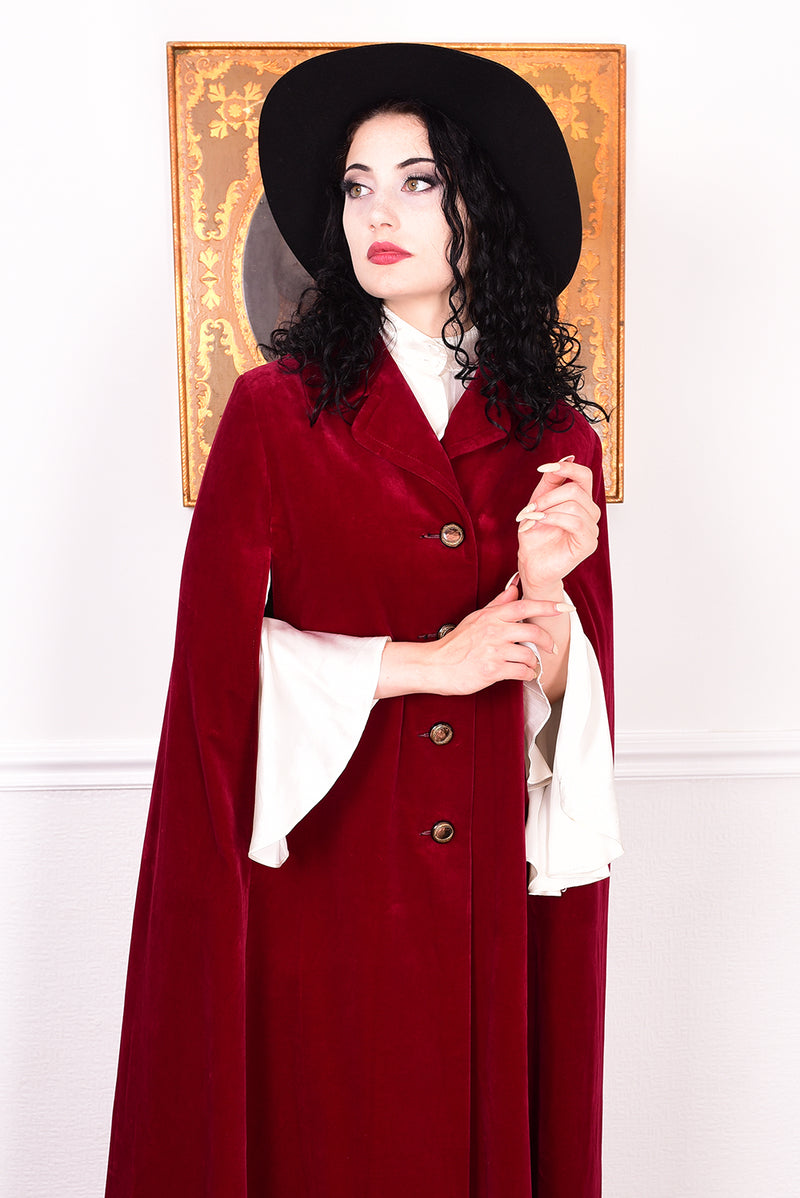 Woman in a vintage 1970s red velvet cape with a black hat standing in a room with a white wall.