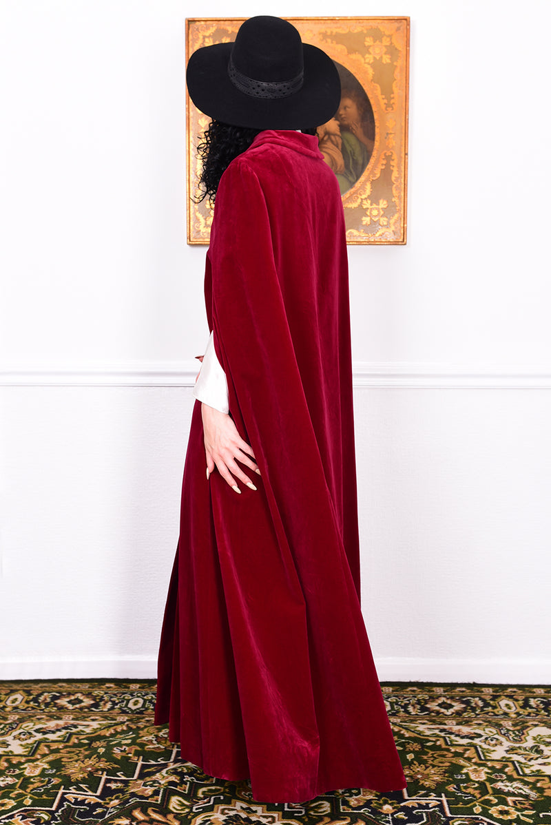 Woman in a vintage 1970s red velvet cape with a black hat standing in a room with a white wall.