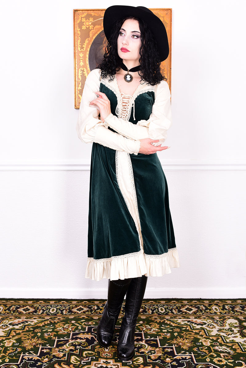 Woman in a vintage 1970s Gunne Sax green velvet dress with white lace details  standing against a white wall.
