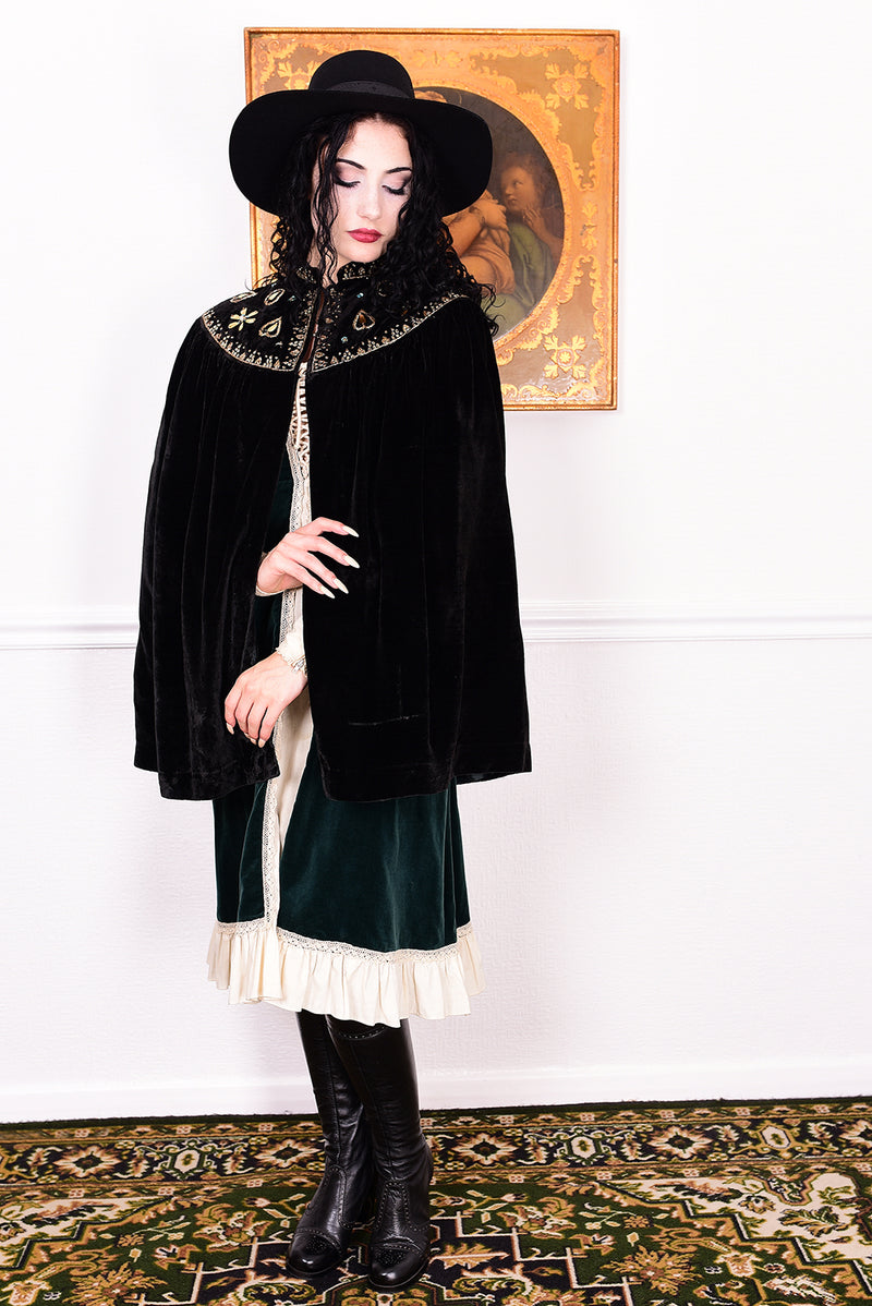 Woman in an antique black velvet cape with embroidered detailing and black boots standing in a room with a white wall.