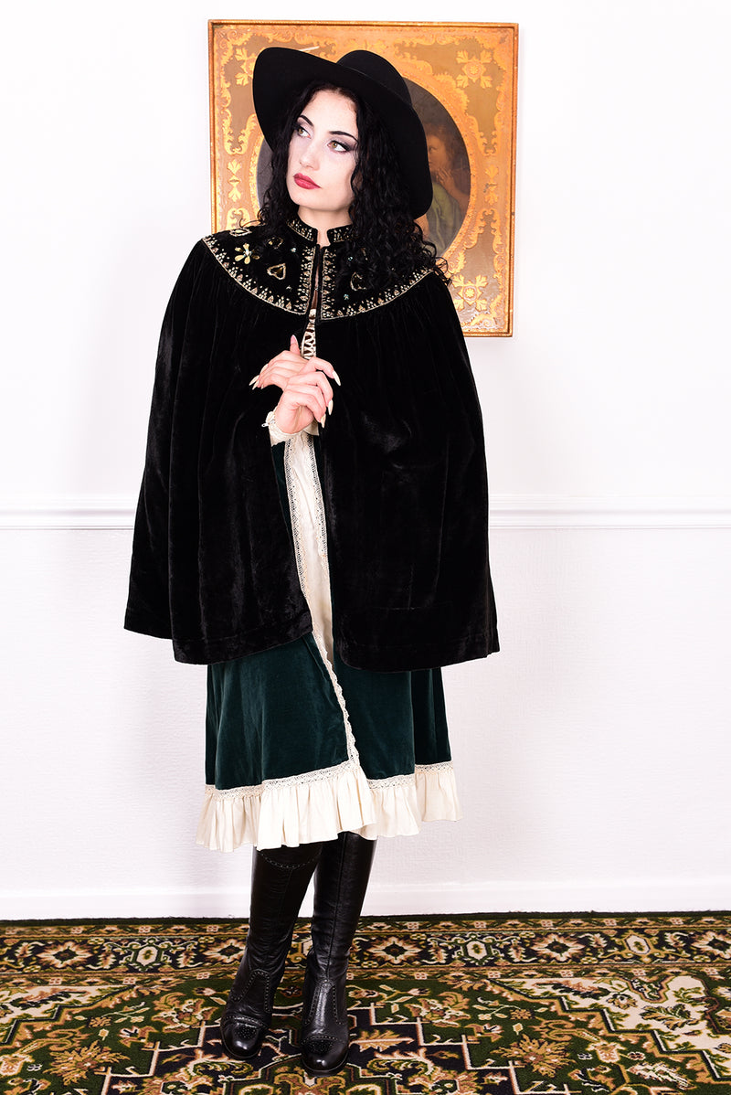 Woman in an antique black velvet cape with embroidered detailing and black boots standing in a room with a white wall.