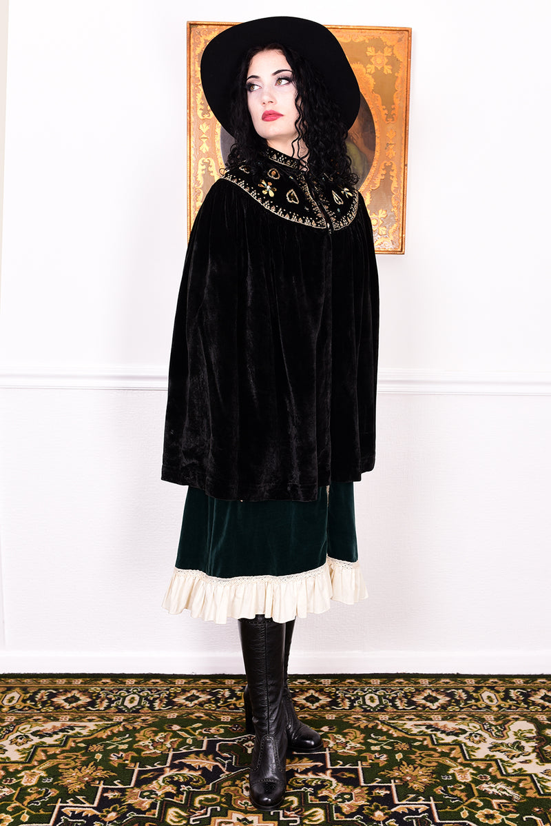 Woman in an antique black velvet cape with embroidered detailing and black boots standing in a room with a white wall.