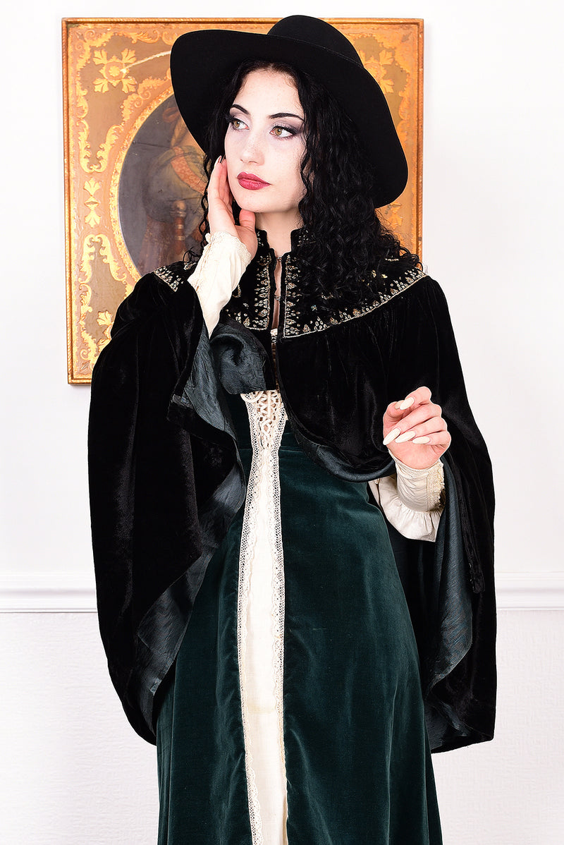 Woman in an antique black velvet cape with embroidered detailing and black boots standing in a room with a white wall.