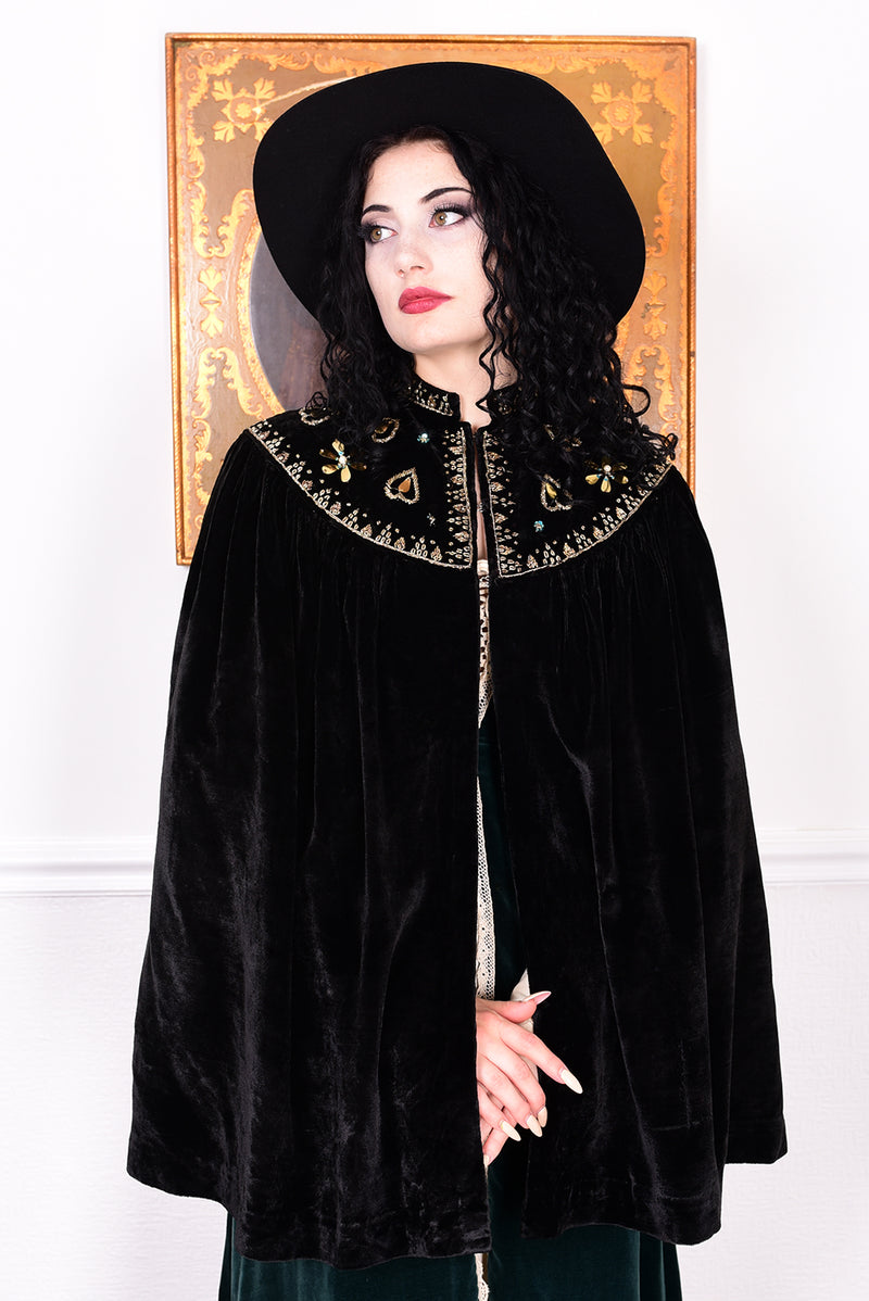 Woman in an antique black velvet cape with embroidered detailing and black boots standing in a room with a white wall.