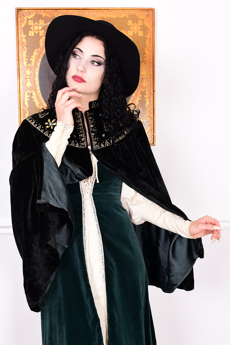 Woman in an antique black velvet cape with embroidered detailing and black boots standing in a room with a white wall.