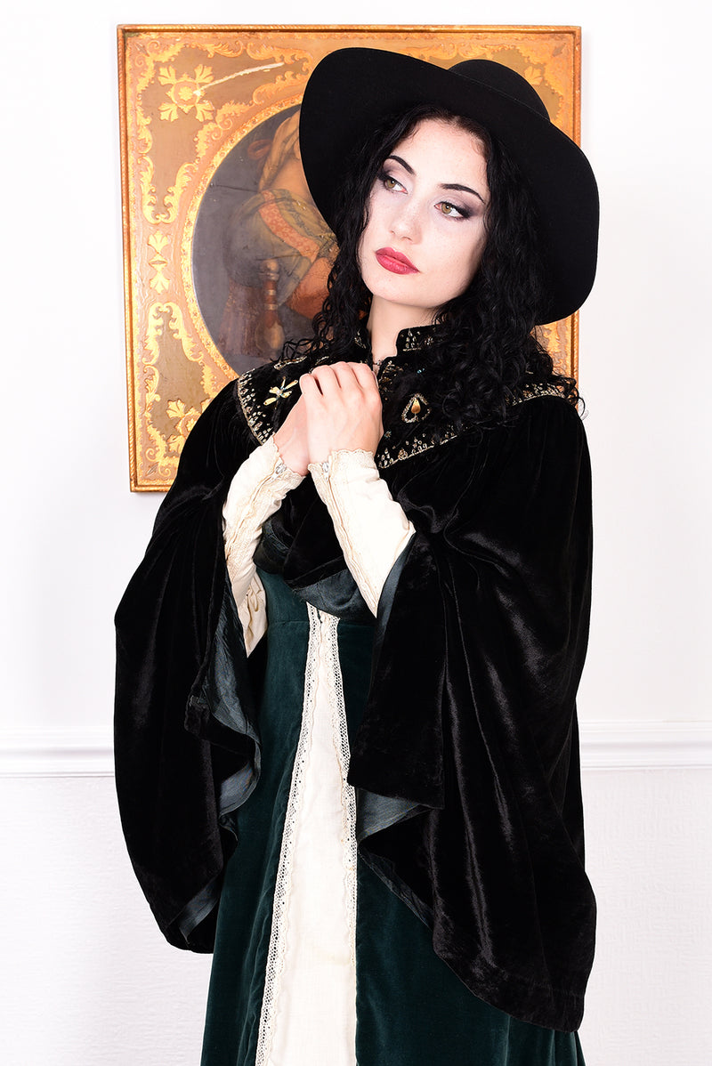 Woman in an antique black velvet cape with embroidered detailing and black boots standing in a room with a white wall.