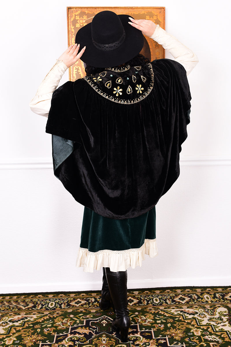 Woman in an antique black velvet cape with embroidered detailing and black boots standing in a room with a white wall.