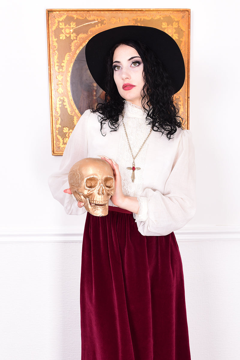 Woman wearing a vintage 1970s maxi dress in white and burgundy velvet by Angela Gore holding a gold skull.