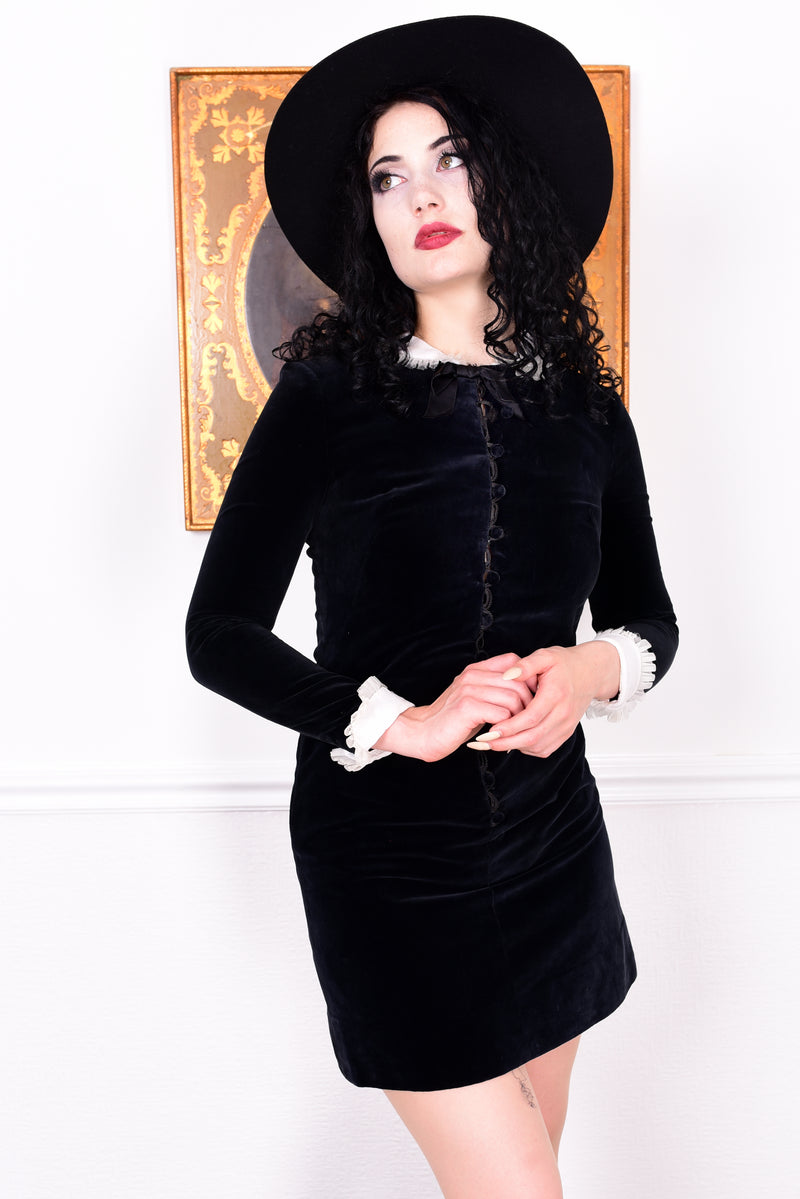 Woman wearing a black vintage 1960s velvet dress and hat holding a book, with a white wall.