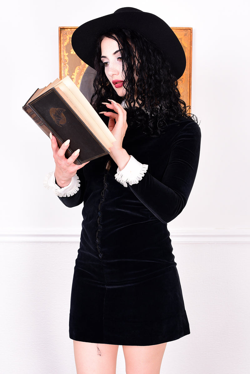 Woman wearing a black vintage 1960s velvet dress and hat holding a book, with a white wall.