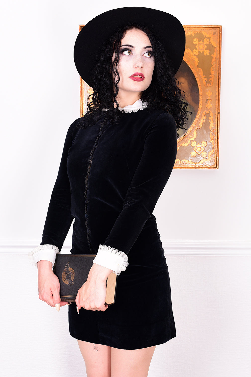Woman wearing a black vintage 1960s velvet dress and hat holding a book, with a white wall.