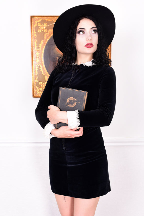 Woman wearing a black vintage 1960s velvet dress and hat holding a book, with a white wall.