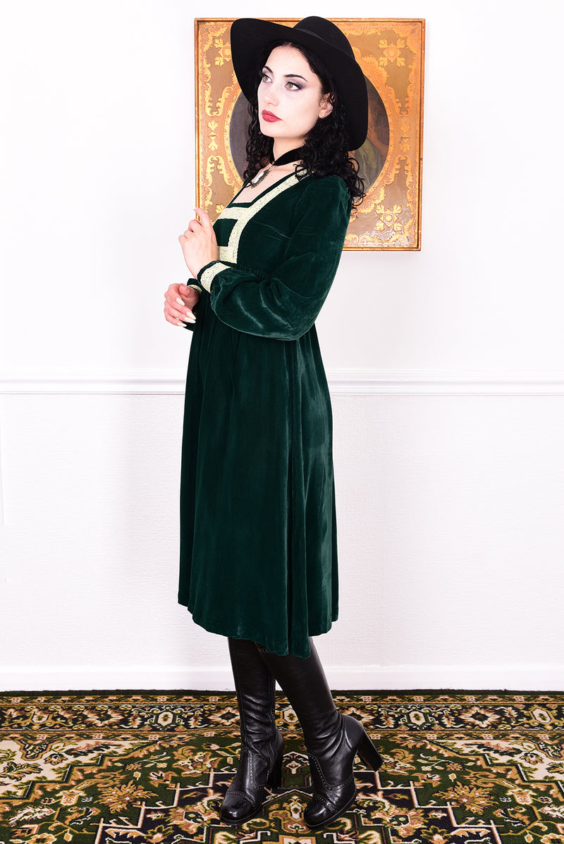 Woman in a vintage 1970s emerald green velvet dress with white lace trim standing against a white wall.