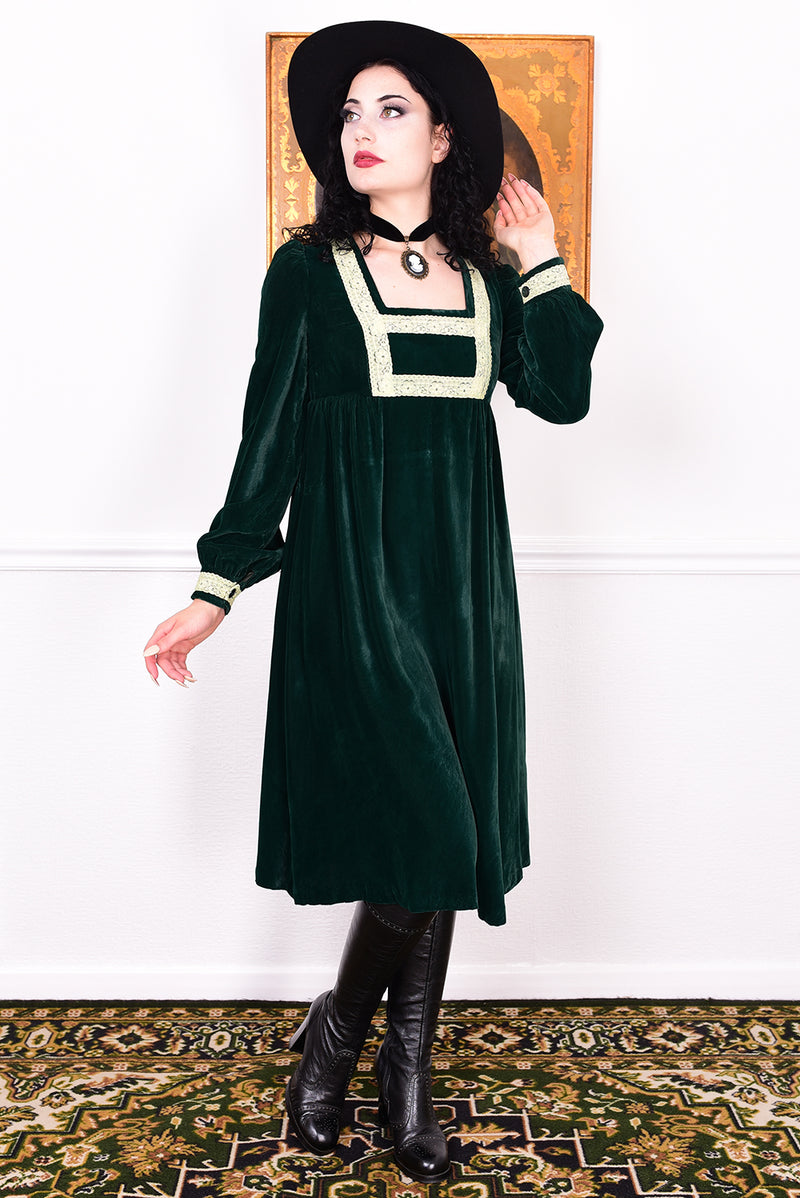 Woman in a vintage 1970s emerald green velvet dress with white lace trim standing against a white wall.