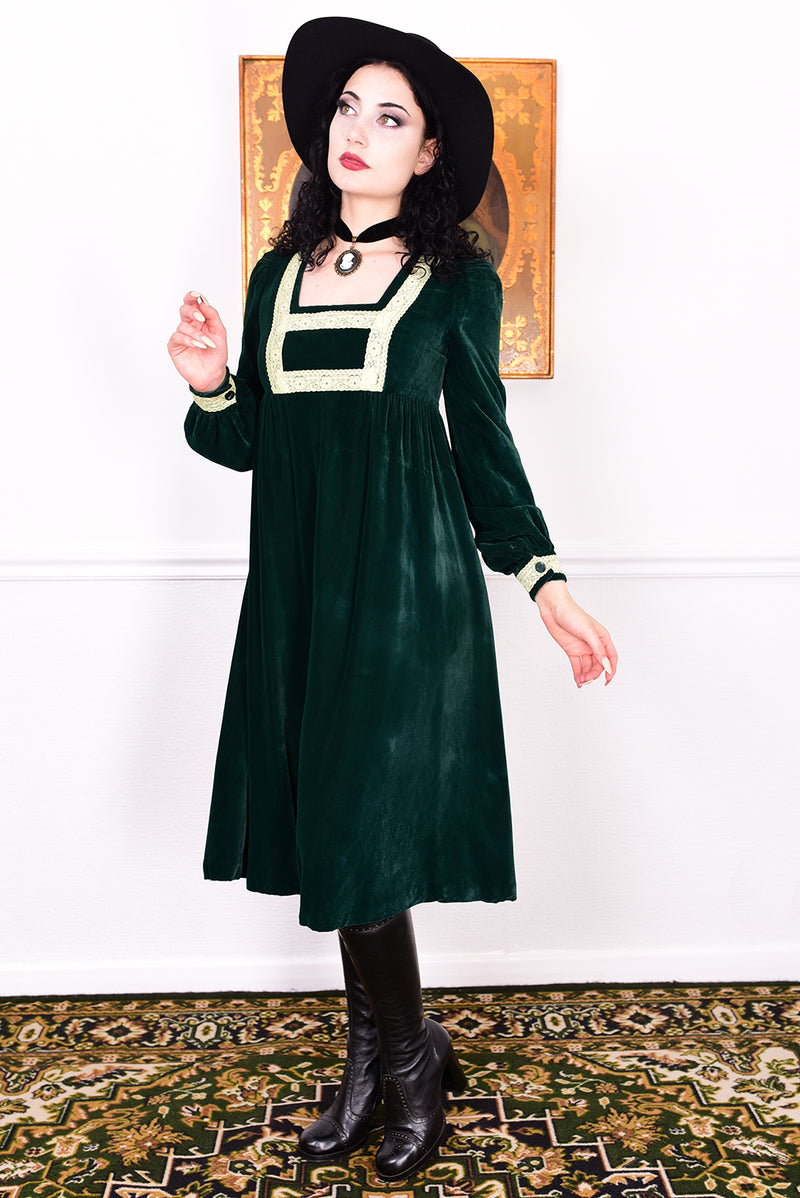 Woman in a vintage 1970s emerald green velvet dress with white lace trim standing against a white wall.