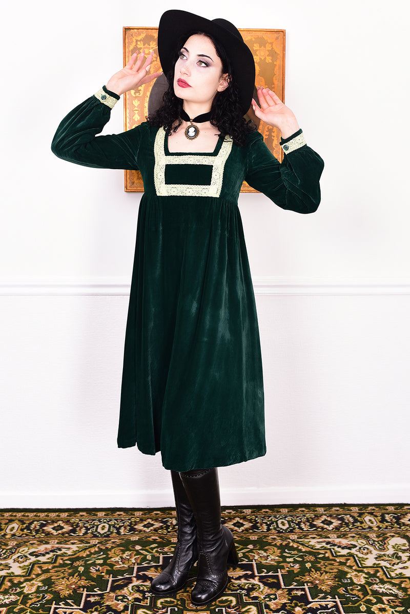 Woman in a vintage 1970s emerald green velvet dress with white lace trim standing against a white wall.