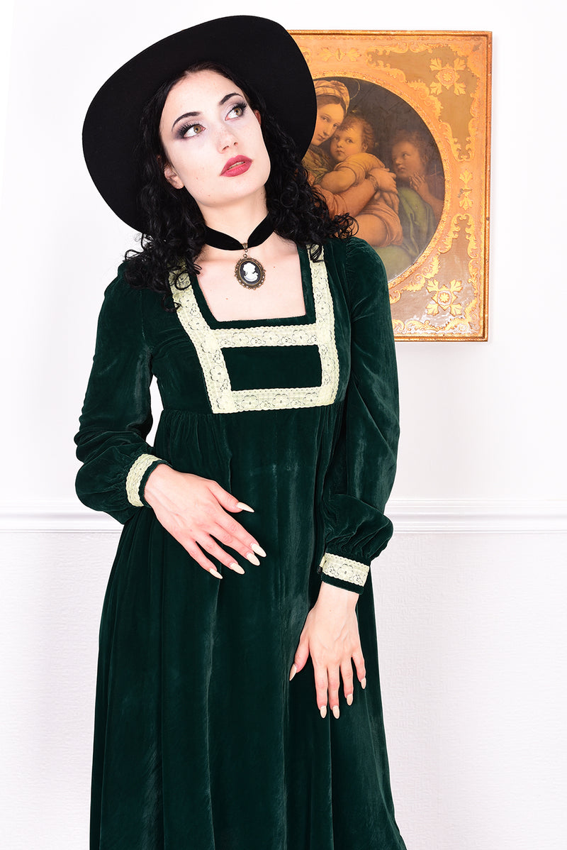Woman in a vintage 1970s emerald green velvet dress with white lace trim standing against a white wall.