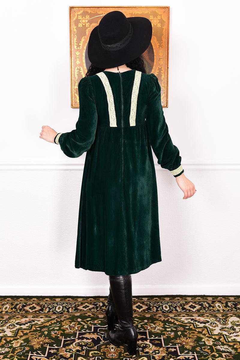 Woman in a vintage 1970s emerald green velvet dress with white lace trim standing against a white wall.