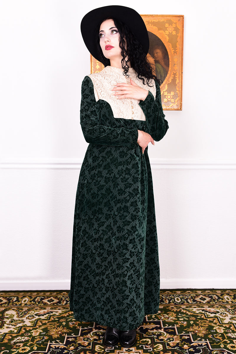 Woman in a vintage 1970s dark green floral print velvet maxi dress with lace details and a black hat.