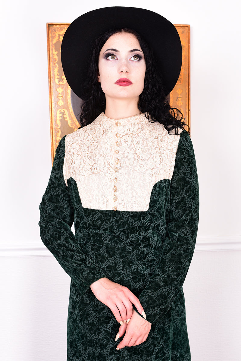 Woman in a vintage 1970s dark green floral print velvet maxi dress with lace details and a black hat.