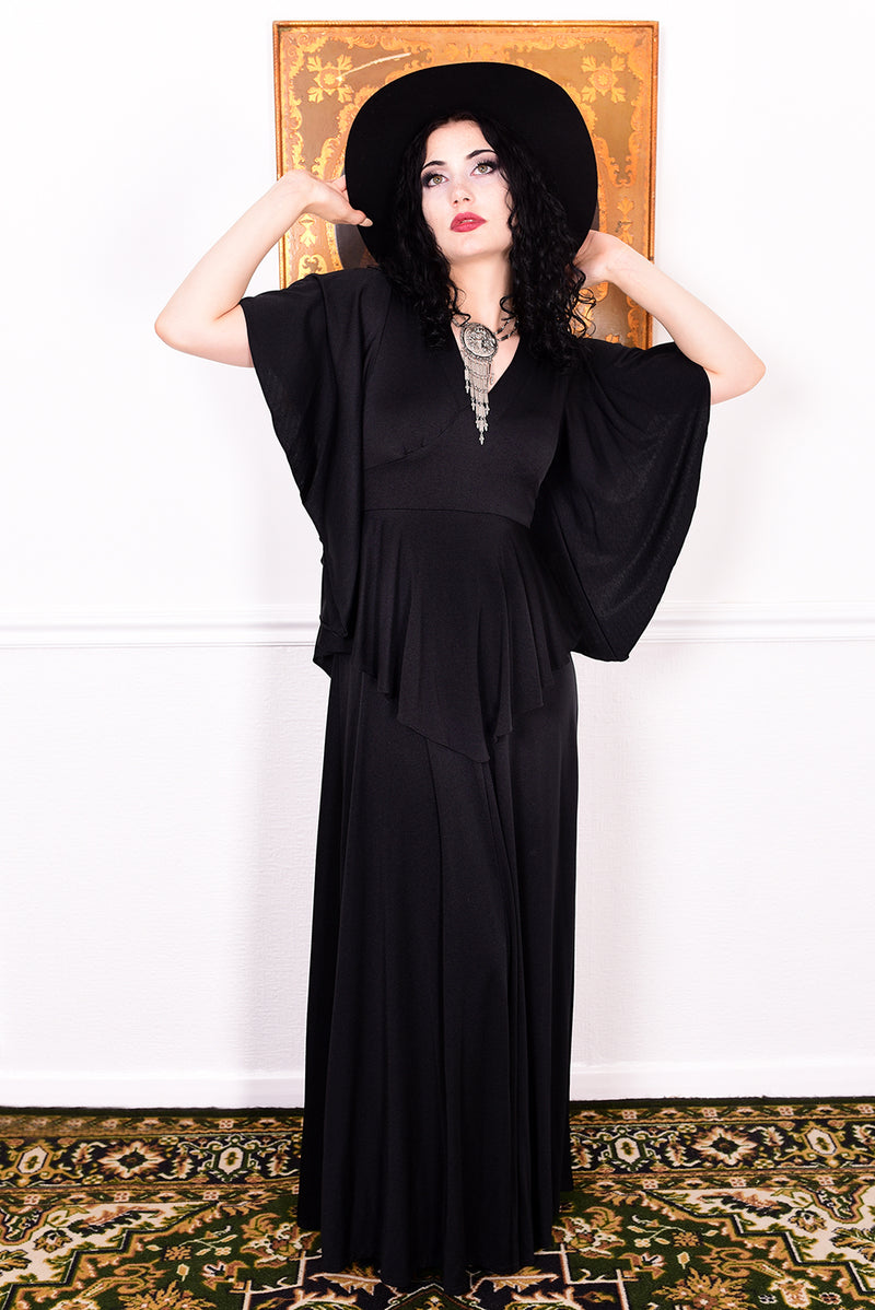 Woman in a black vintage 1970s gothic dress and hat posing against a white wall.