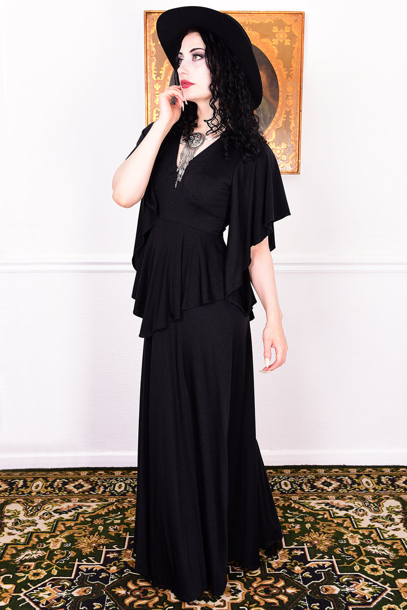 Woman in a black vintage 1970s gothic dress and hat posing against a white wall.