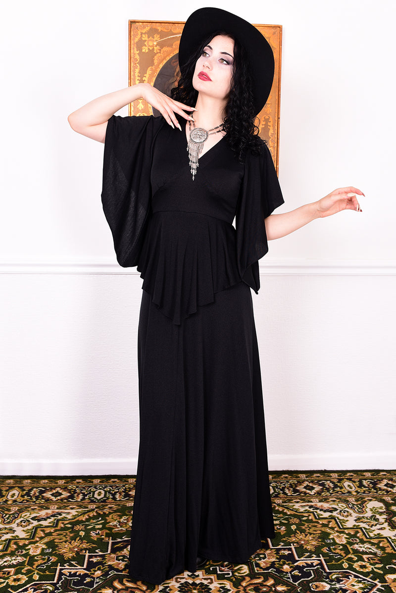 Woman in a black vintage 1970s gothic dress and hat posing against a white wall.