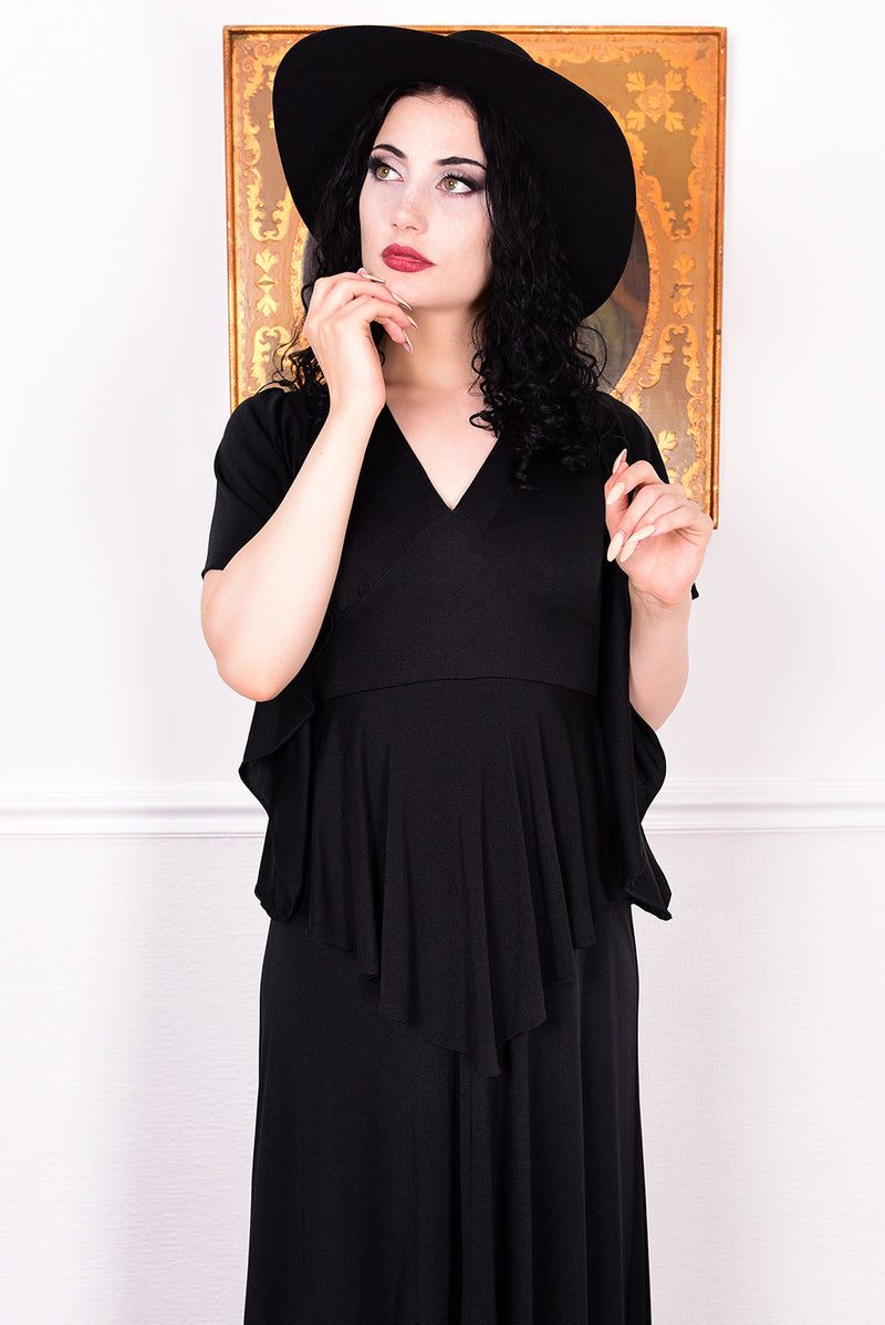 Woman in a black vintage 1970s gothic dress and hat posing against a white wall.
