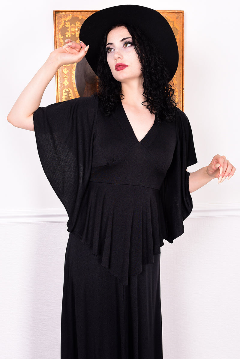 Woman in a black vintage 1970s gothic dress and hat posing against a white wall.