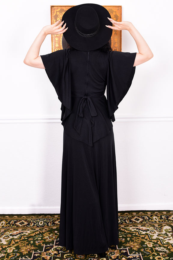 Woman in a black vintage 1970s gothic dress and hat posing against a white wall.