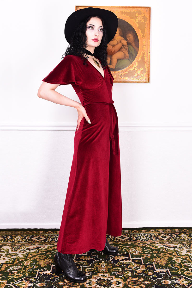 Woman in a red vintage velvet jumpsuit and black hat posing in a room with a painting on the wall.