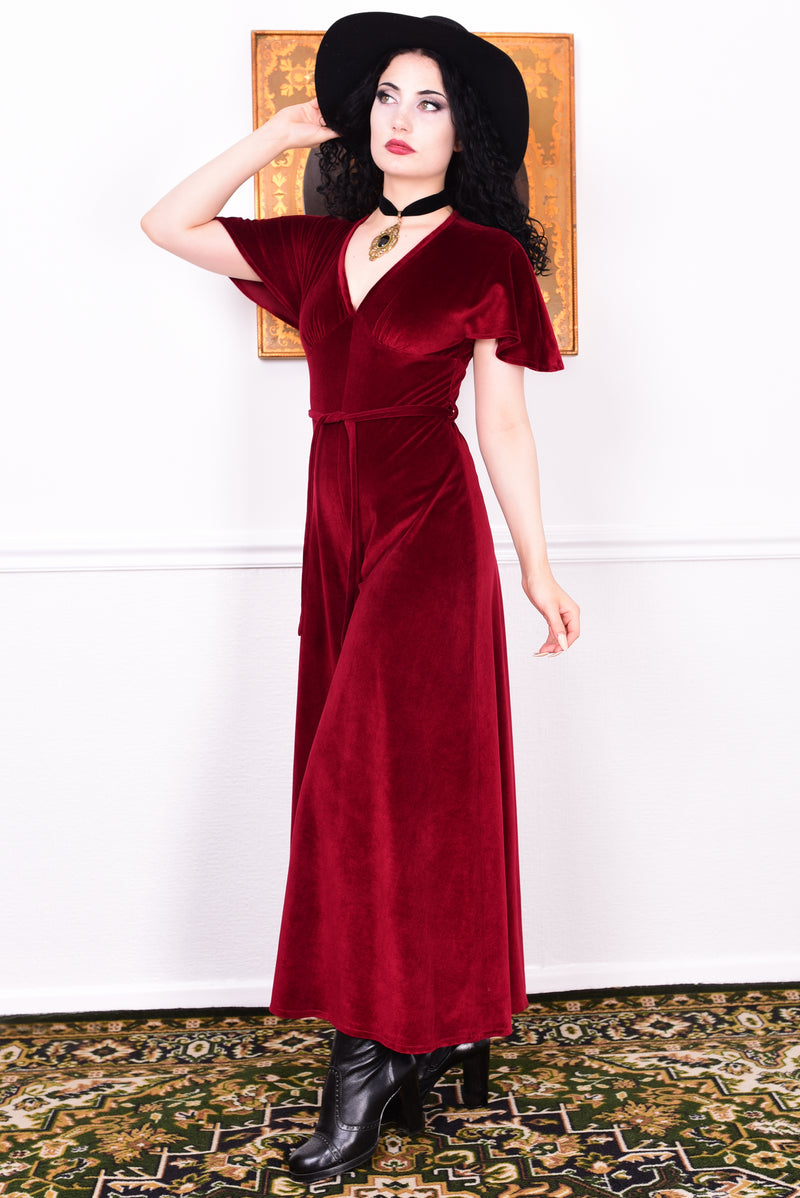 Woman wearing a red vintage velvet jumpsuit dress with a black hat and boots, standing against a white wall.