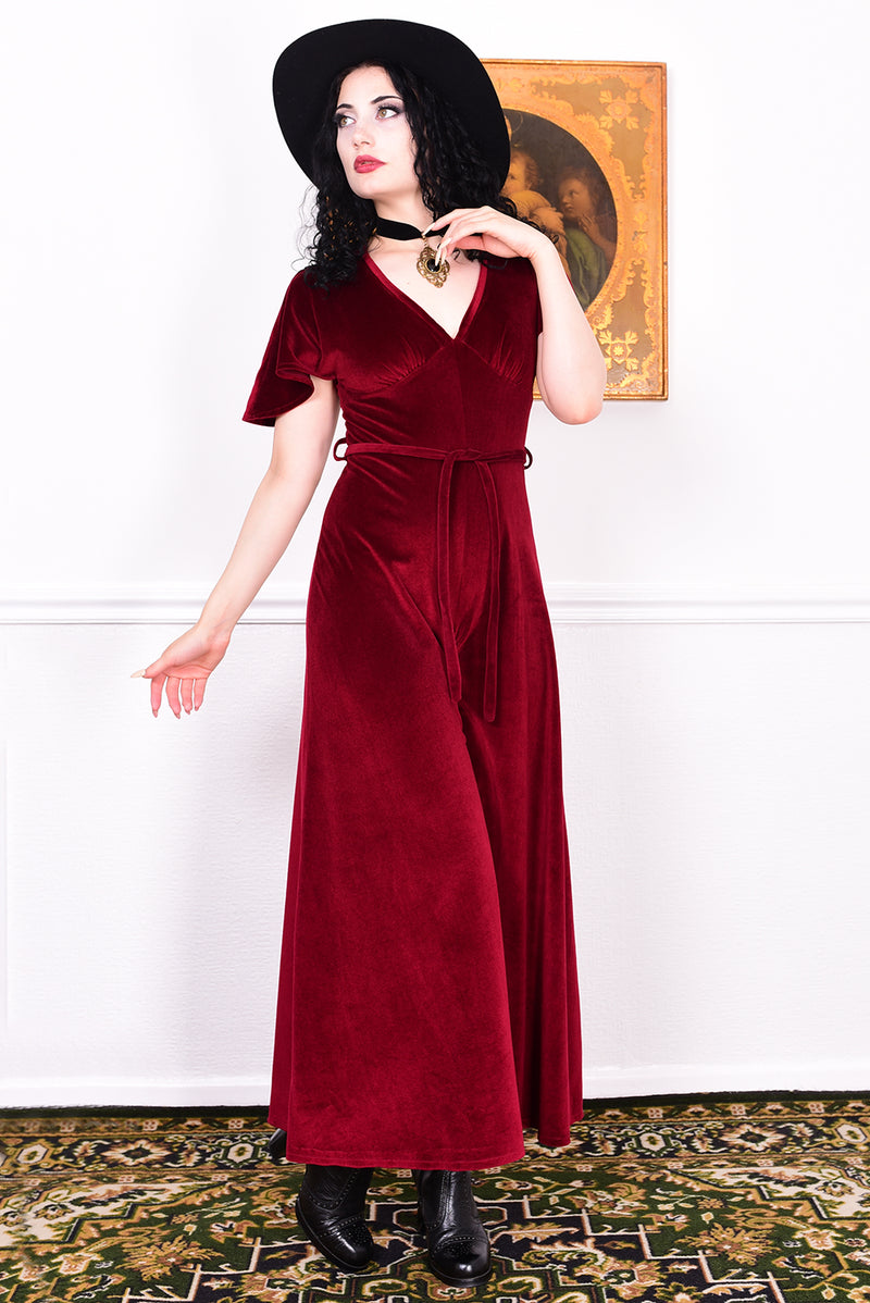 Woman in a red vintage velvet jumpsuit and black hat standing against a white wall with a decorative rug.
