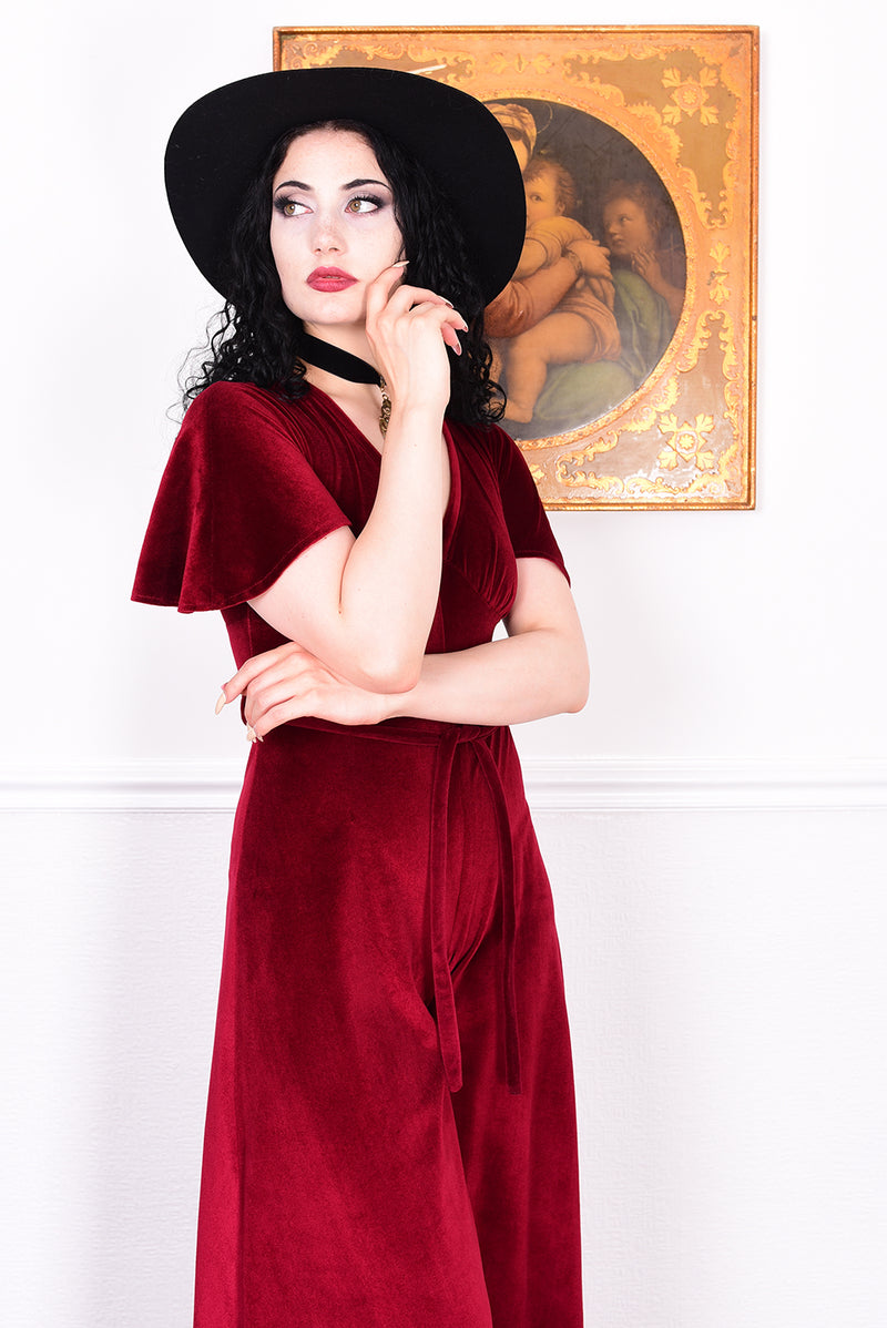 Woman in a red jumpsuit and black hat posing in front of a decorative painting.