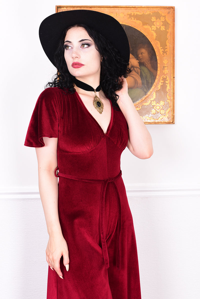 Woman wearing a red velvet jumpsuit and black hat indoors with a decorative painting in the background.