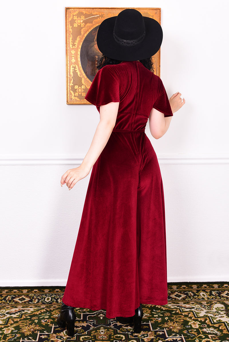 Woman wearing a burgundy velvet jumpsuit and black hat in a room with a decorative rug and wall art.