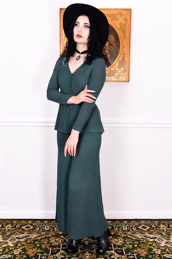 Woman in a vintage 1970s green trouser suit and black hat standing in a room with white wall.