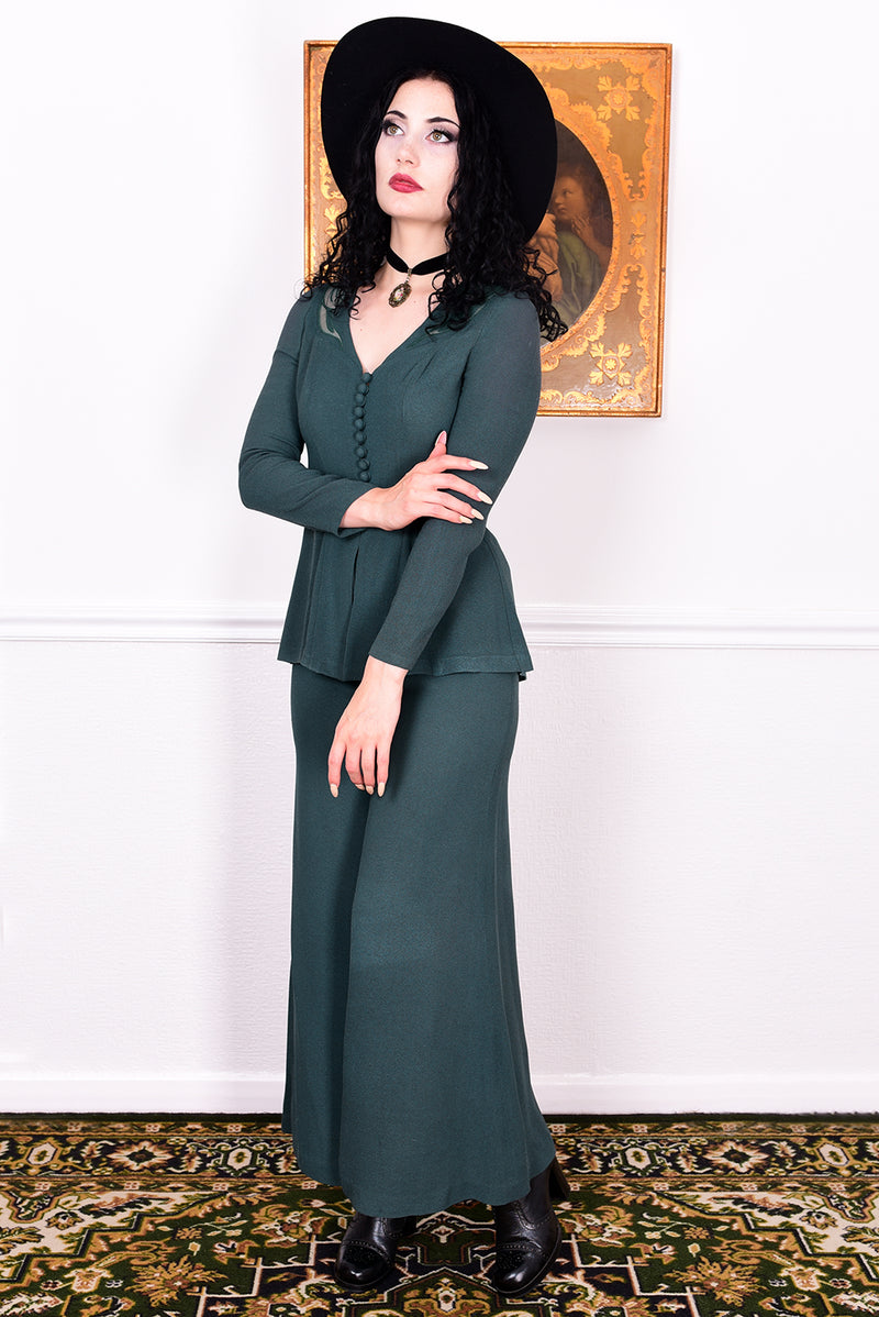 Woman in a vintage 1970s green trouser suit and black hat standing in a room with white wall.