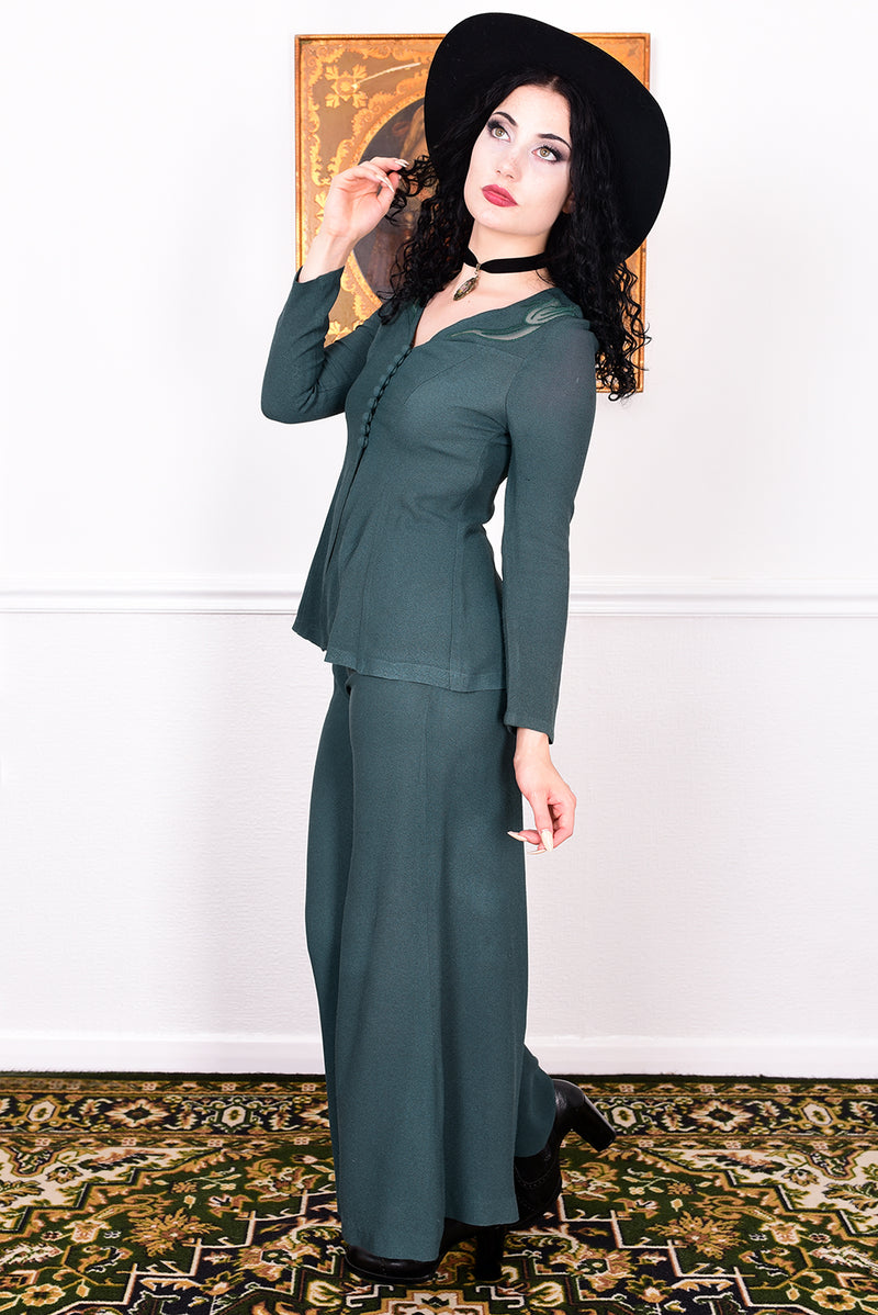 Woman in a vintage 1970s green trouser suit and black hat standing in a room with white wall.